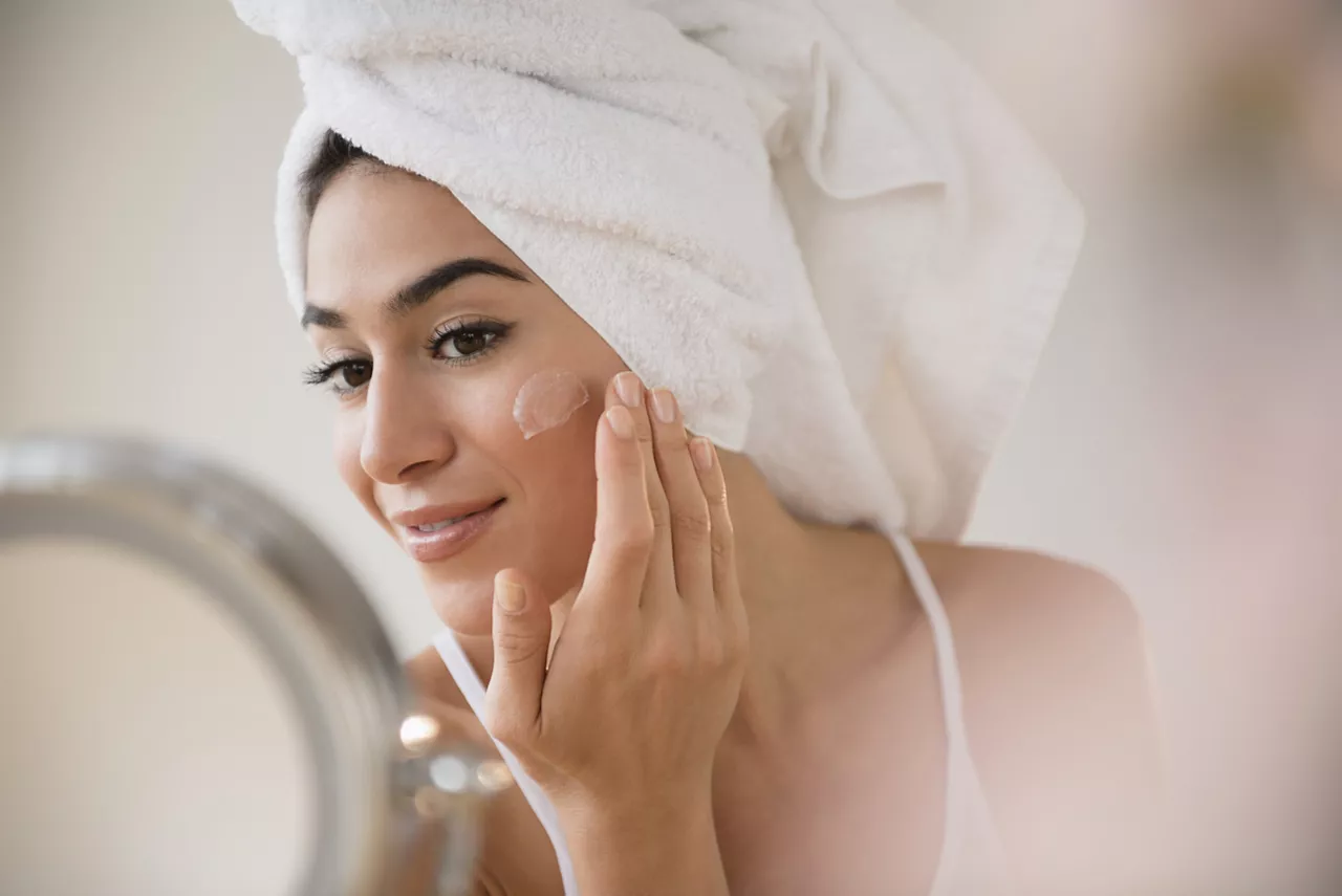 Woman with hair in towel rubbing lotion on face in front of mirror.