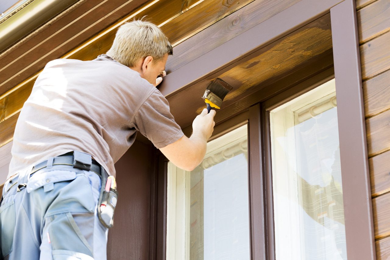 Contractor with paintbrush painting wooden house exterior.