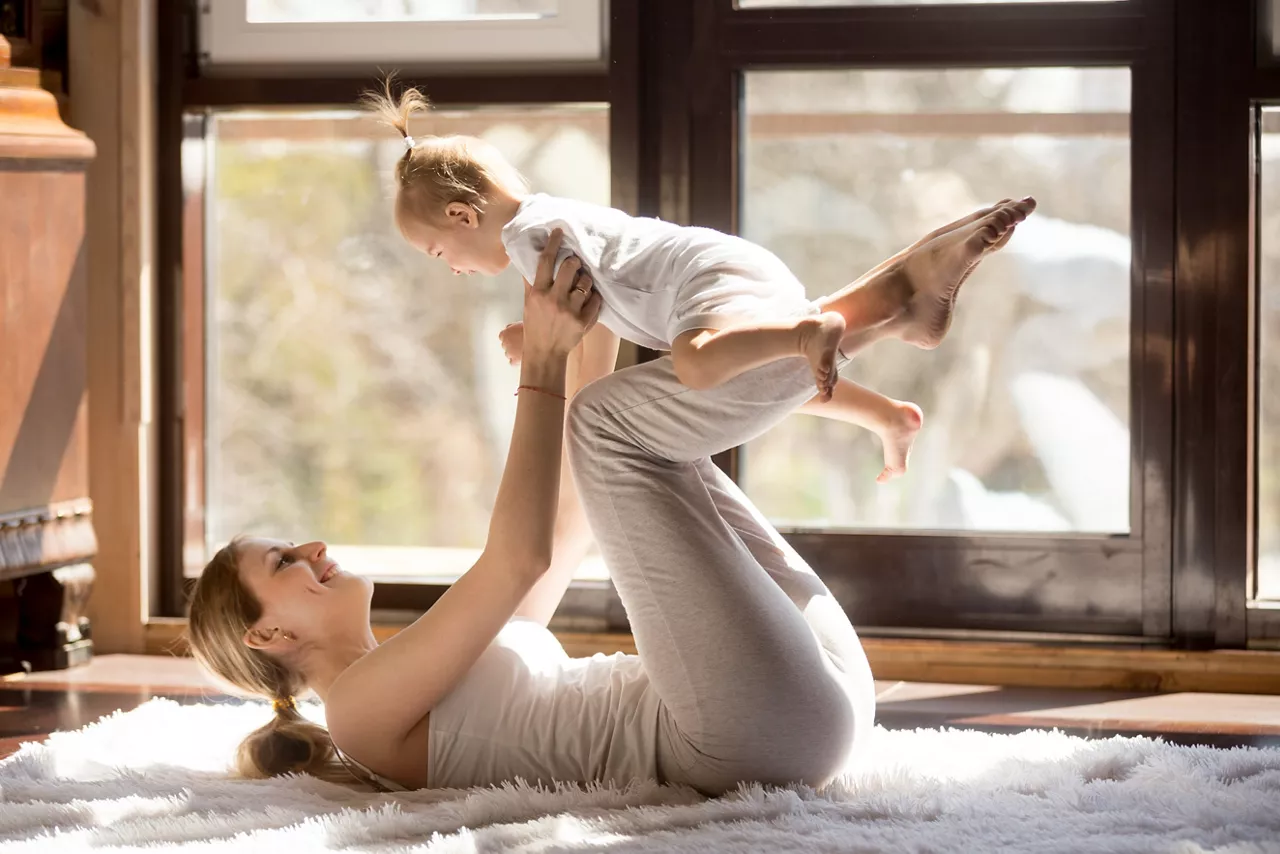 Young mother working out wearing white sportswear,  exercising at home with baby daughter, leg lifting with kid as a weigh, exercising and bonding with child, enjoyment. Healthy lifestyle concept