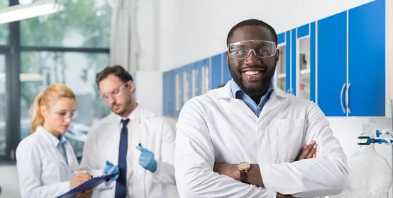 Happy Smiling African American Scientist Stand In Front Of Colleagues In Laboratory Making Notes Of Experiment Or Research Results, Mix Race Team In Modern Science Lab