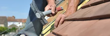 Man working on roof shingles with a hammer.