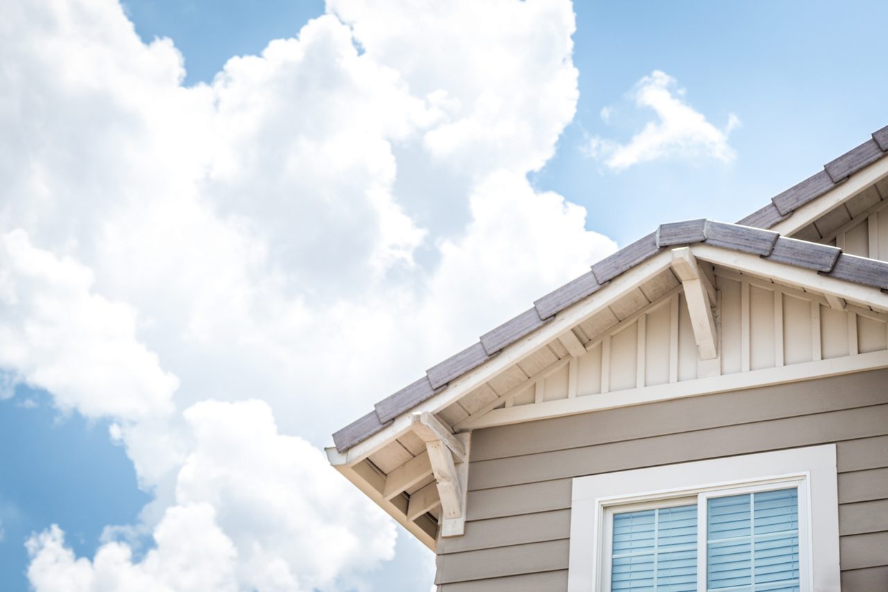 Canto da casa com janela e telhado com nuvens brancas e céu azul ao fundo