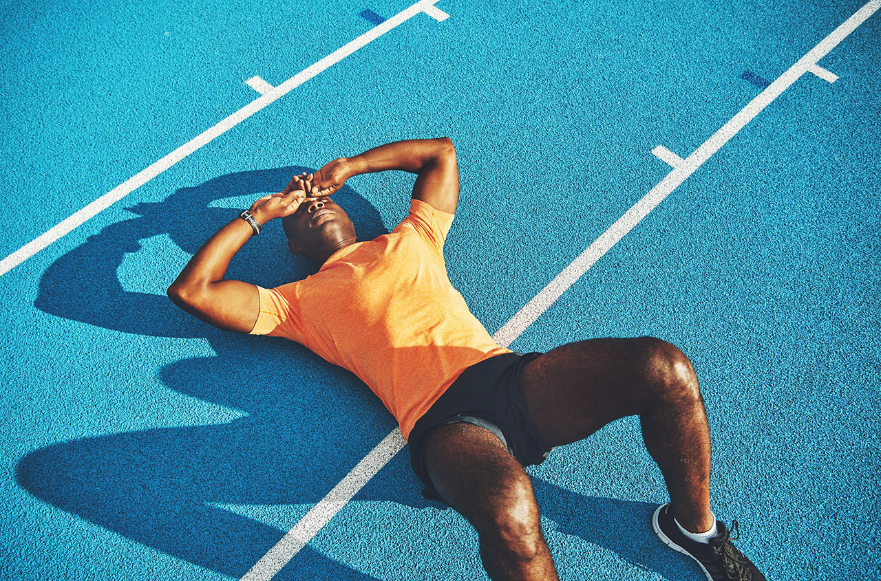 Exhausted young athletic African man in sportswear lying on the lanes of a running track taking a break from training