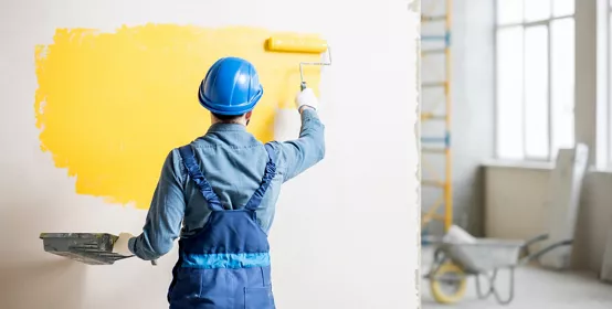 Workman in uniform painting wall with yellow paint at the construction site indoors