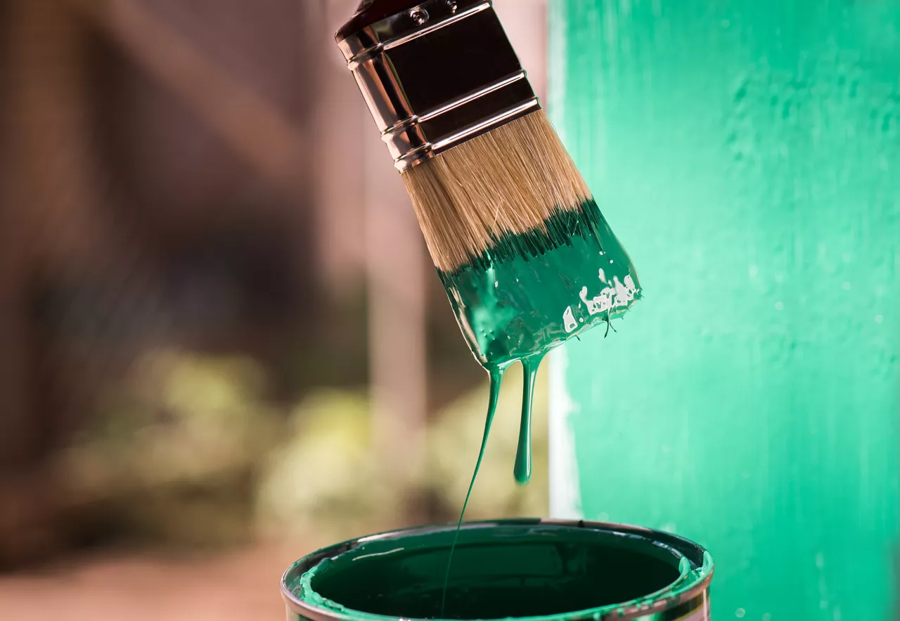 Paint brush dipping out of can of green paint with freshly painted wall in background