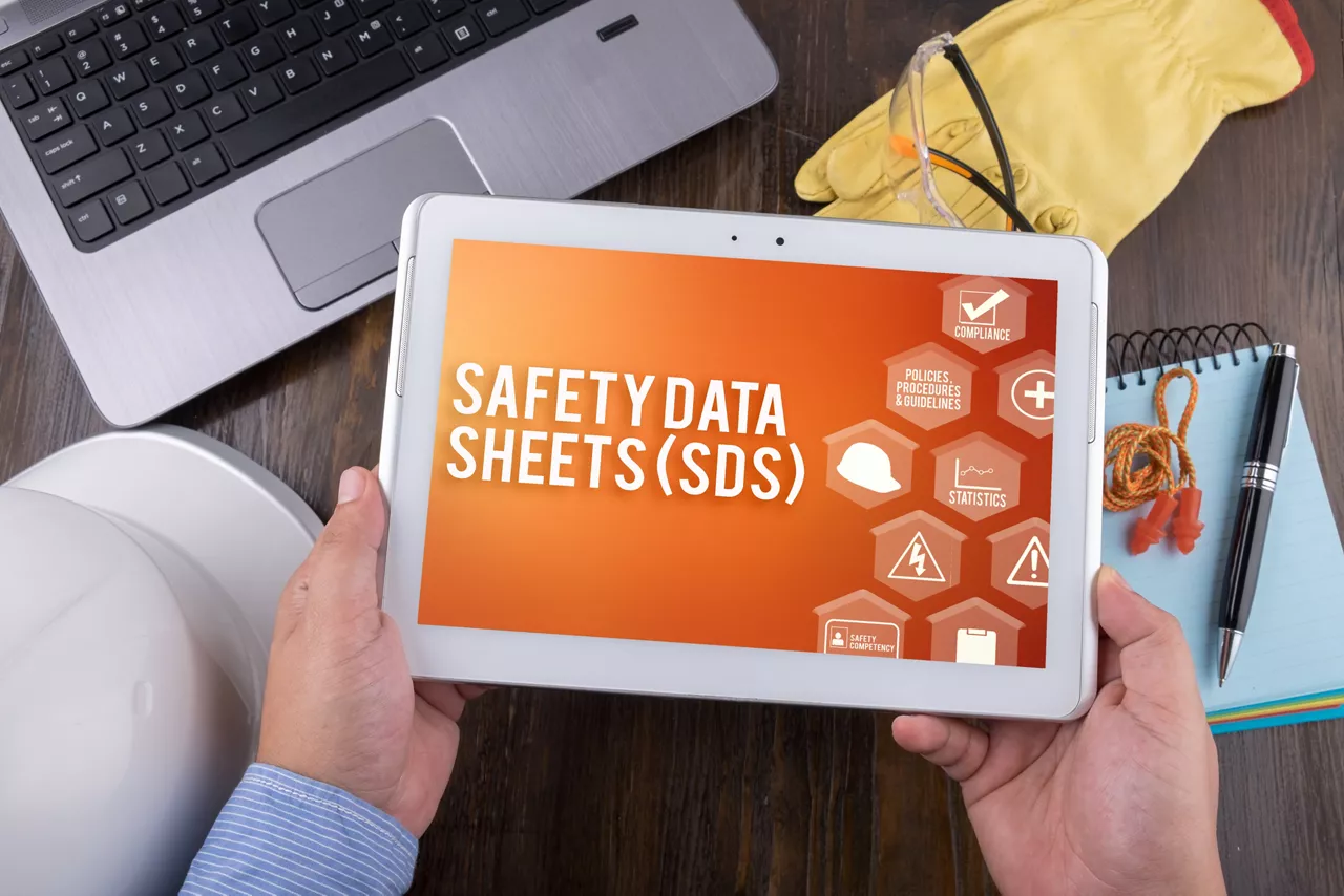 Hands holding tablet showing graphic for Safety Data Sheets, desk background.