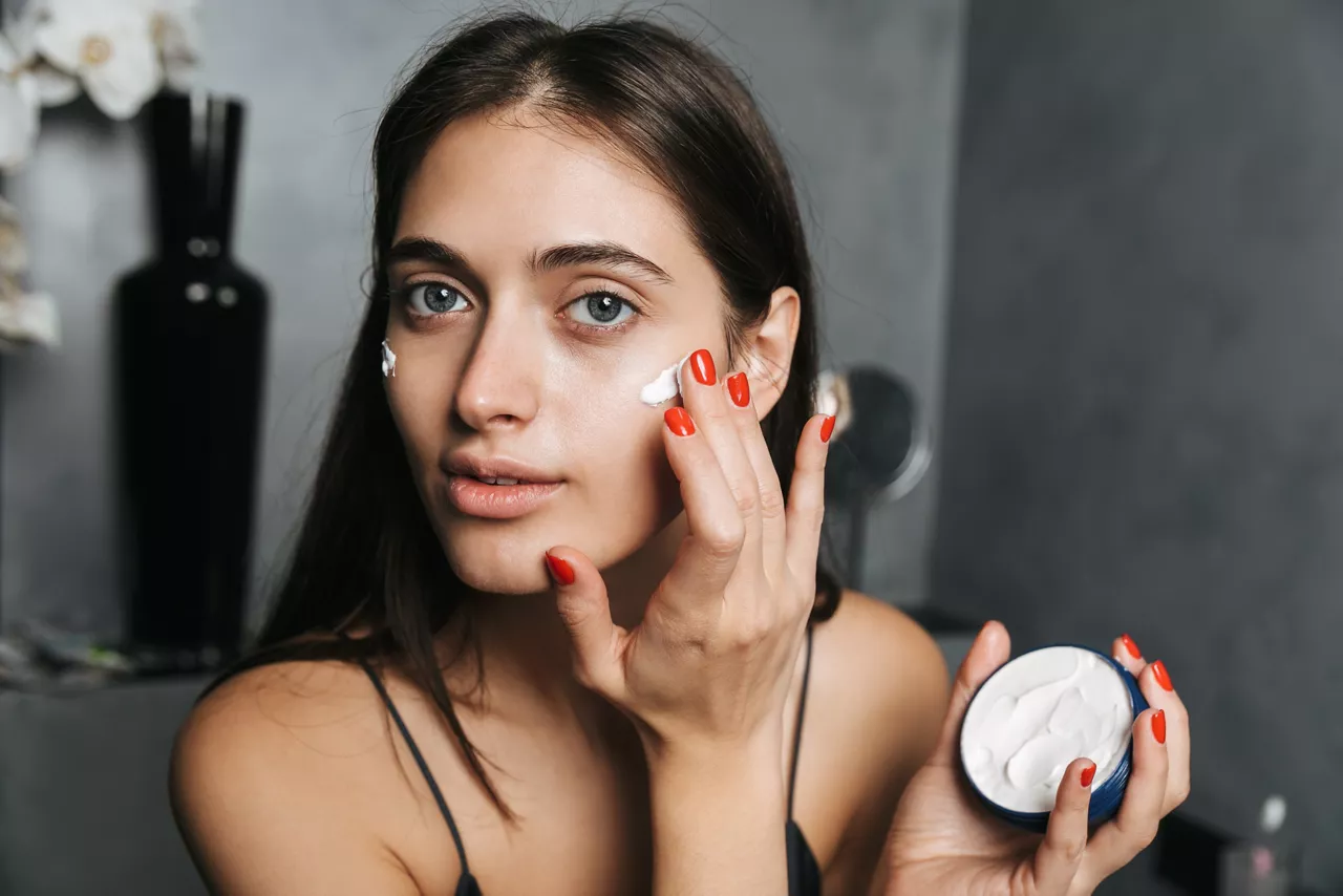 Photo of pretty woman with long dark hair standing in bathroom and applying moisturizing cream on body