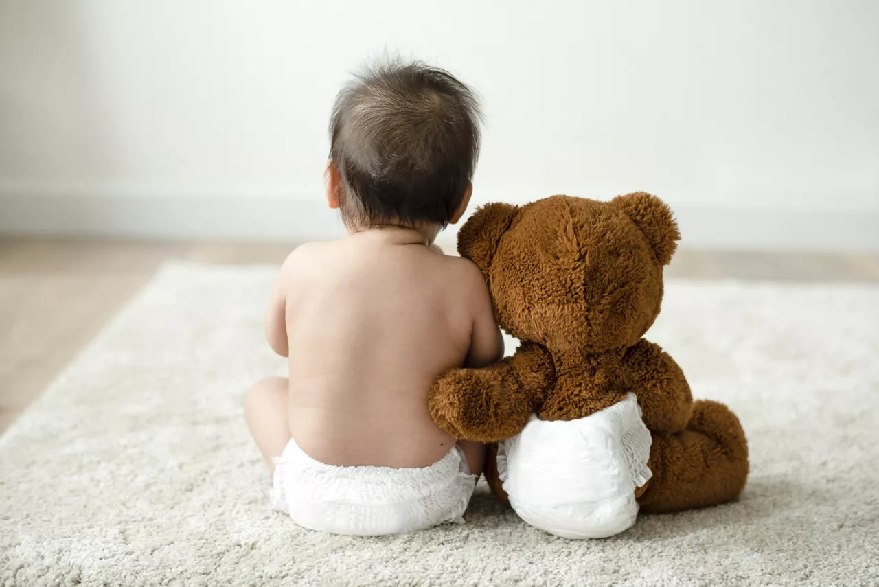 Back of a baby with a teddy bear, both are dressed in diapers