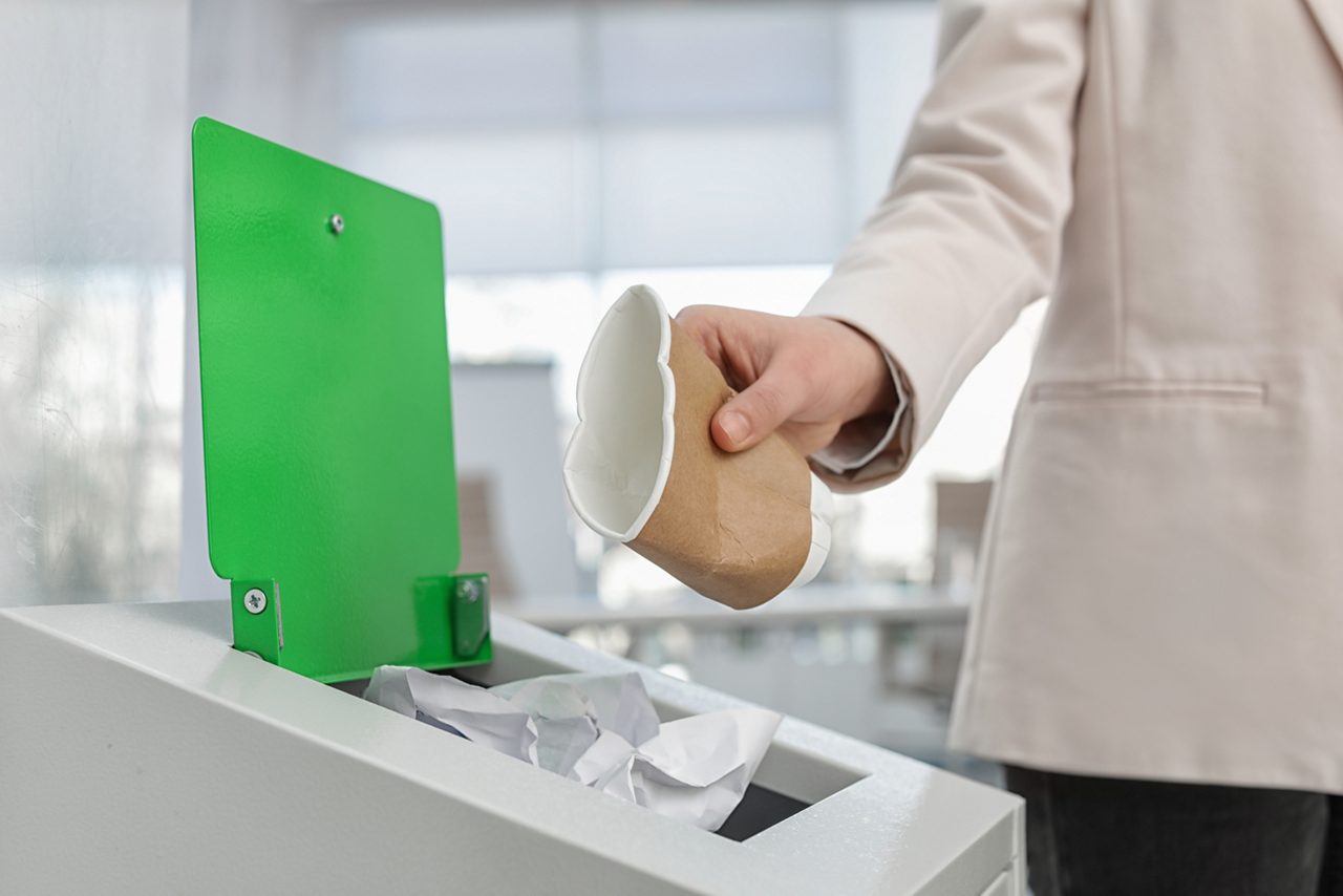 Woman putting used paper cup into trash bin in modern office, closeup. Waste recycling