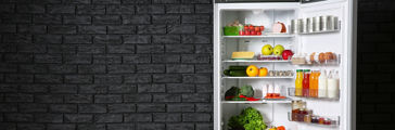 Tall stainless steel refrigerator with door open filled with fruits and vegetables on black brick wall background