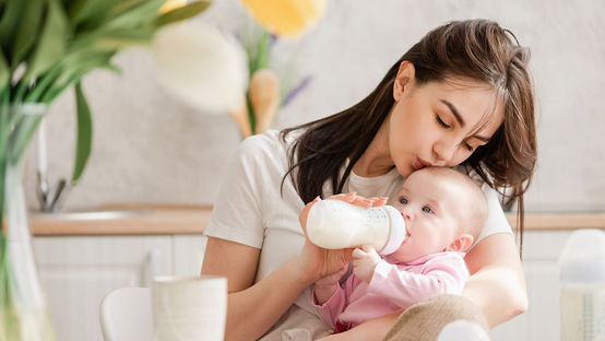 牛乳を飲む時に、若い女性が赤ちゃんにキスをする 赤ちゃんの授乳。新生児に瓶詰めの調合液を摂食する。