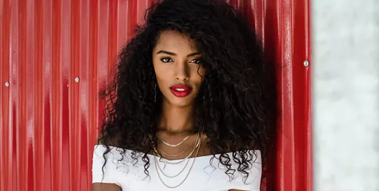 Fashionable black woman with red lipstick and natural hair leaning in a red wall posing on the street while looking at camera.