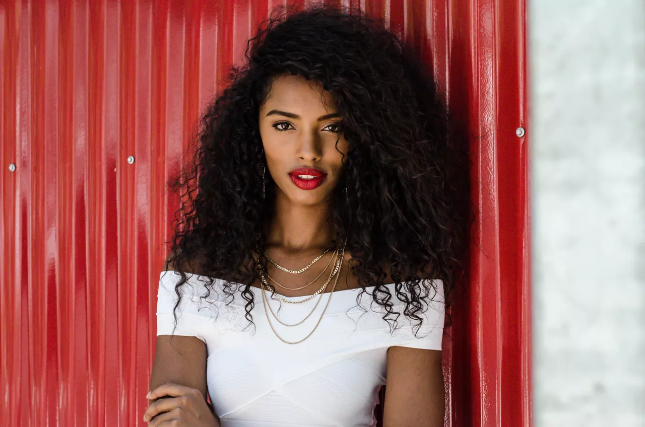 Fashionable black woman with red lipstick and natural hair leaning in a red wall posing on the street while looking at camera.