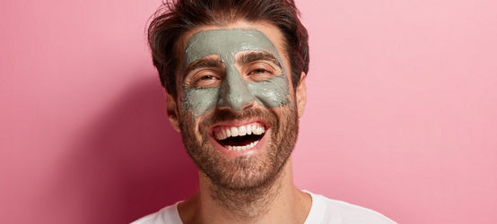 Joyful delighted man has clay mask on face, enjoys spa treatments, has broad smile, being in high spirit, cares about beauty, wears white t shirt, isolated on pink wall. Skin care and wellness concept