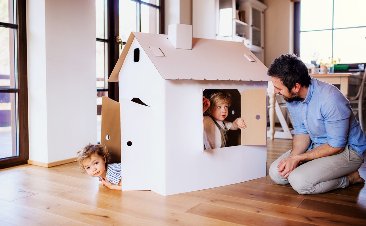 Two happy toddler children with father playing with paper house indoors at home.