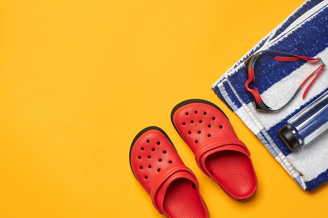 Deitado plano, acessórios de piscina, como óculos de natação, garrafa transparente, toalha listrada e crocodilos vermelhos em um fundo amarelo. Roupa de praia multicolorida. Espaço de cópia, vista superior, close-up.