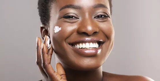 Skin Care Concept. Closeup Portrait Of Happy African American Woman Applying Moisturizing Cream on Face Over Grey Background