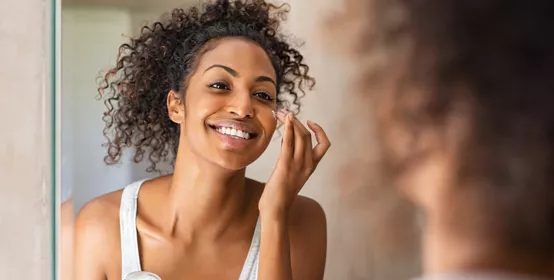 Young african woman applying moisturizer on her face while standing  