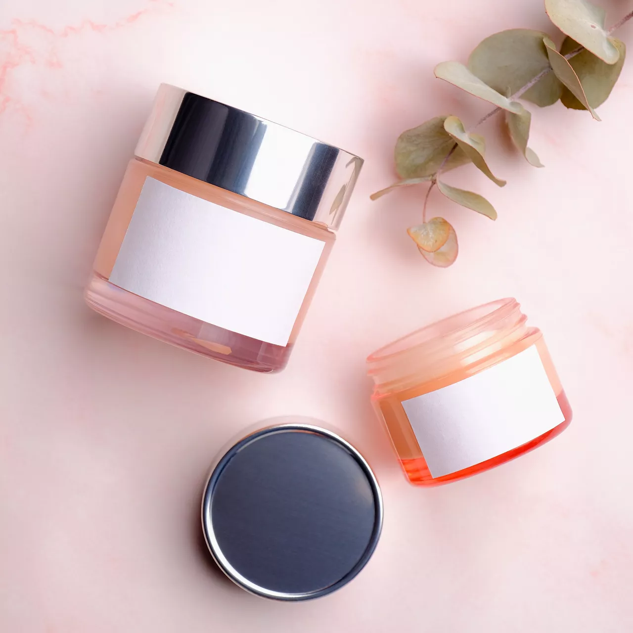 Skincare product packaging mockup. Jars of cosmetic cream with white labels and eucalyptus leaf on pink background. Flat lay, top view.