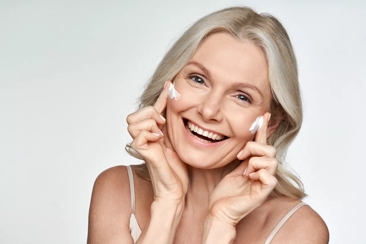 Happy 50s mid aged mature old blonde lady applying facial cream on face skin laughing enjoying anti age fresh soft skin care beauty spa treatment skincare creme isolated on white background. Portrait.