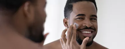 Happy handsome African American man applying moisturizer on face, putting collagen anti age cream, sunscreen, cleansing lotion, caring for hydrating soft skin, reducing wrinkles, acne, eyebags