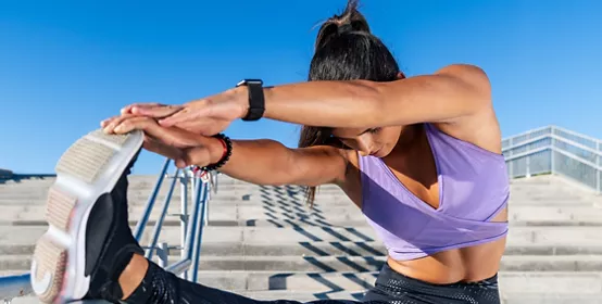 Flexible slender female in sportswear leaning on railing on stairs and stretching legs while warming up and doing forward bend exercise during workout in city.