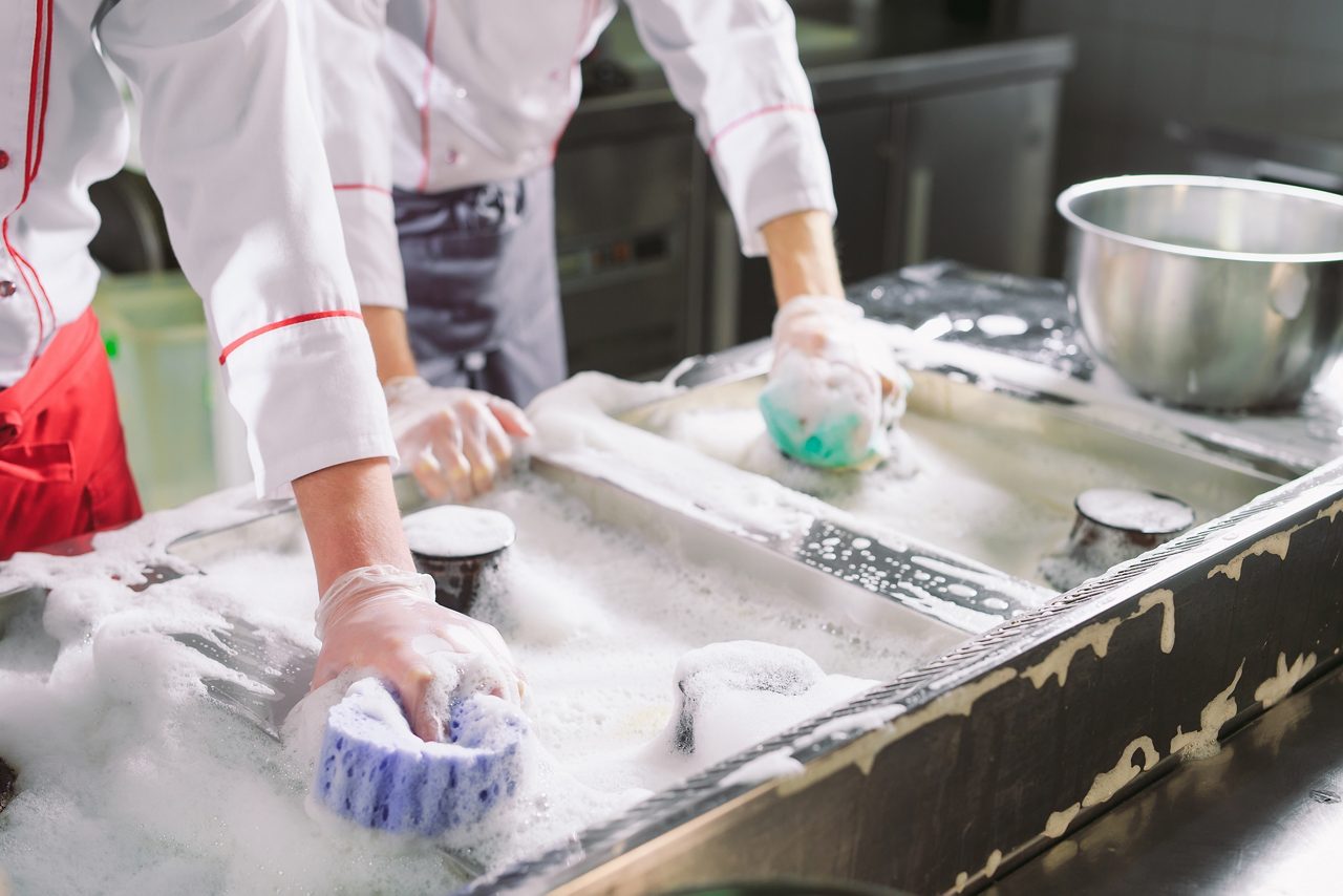 Two restaurant workers scrubbing down kitchen stove with sponges and soapy water