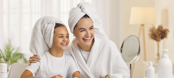 Happy mother and child enjoying morning skin care routine together. Cheerful beautiful mommy and little daughter looking in small mirror on beauty table in bedroom, applying face cream and laughing