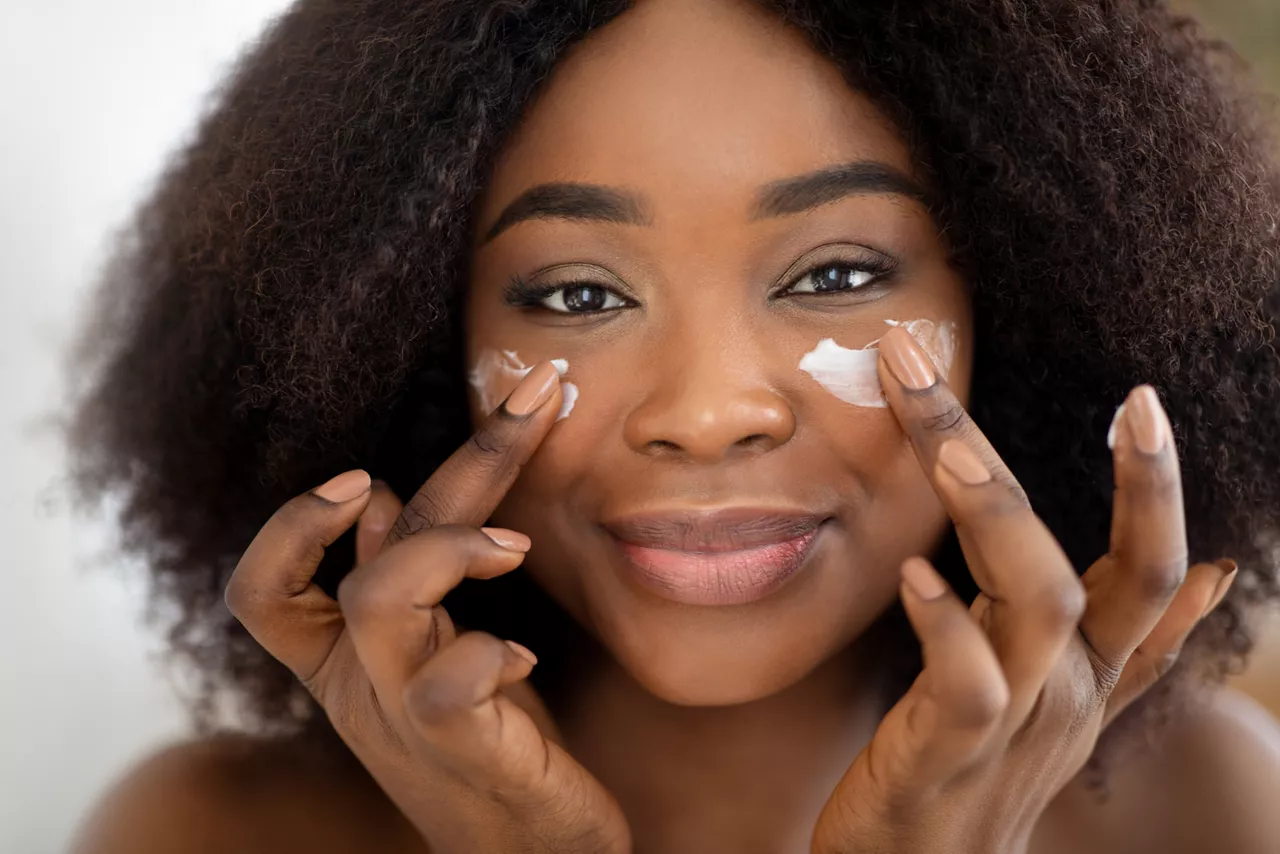 Closeup portrait of attractive millennial black lady applying facial cream under her eyes. Pretty African American woman taking care of skin, putting on nourishing lotion. Beauty and natural cosmetics