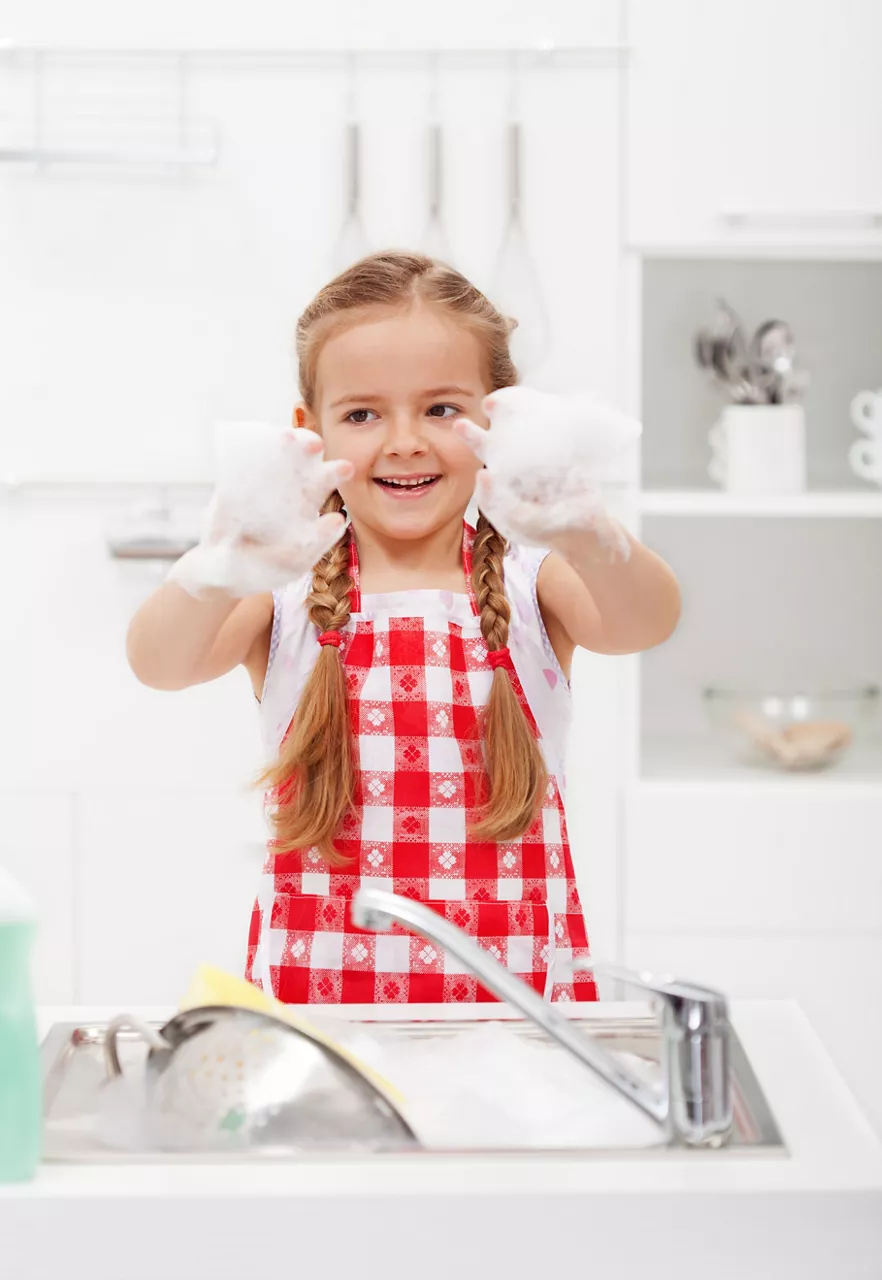 Happy little girl playing with bubbles from the kitchen sink full of dishes 