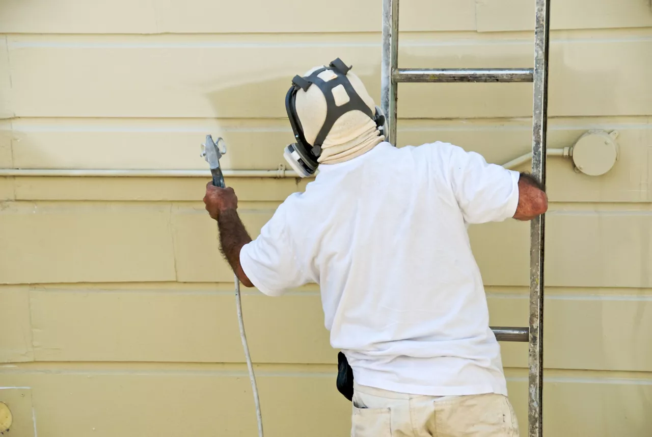 A man on a ladder uses spray gun to paint the exterior of a building 