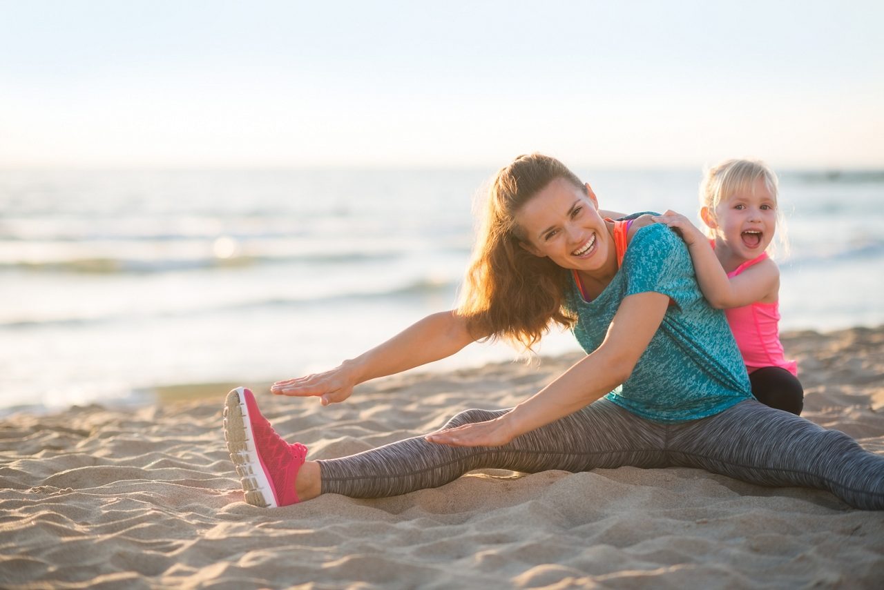 Healthy mother and baby girl stretching on beach in the evening