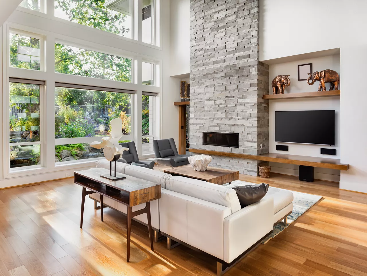 Beautiful living room with hardwood floors, fireplace, and large bank of windows with view of lush vegetation, in new luxury home