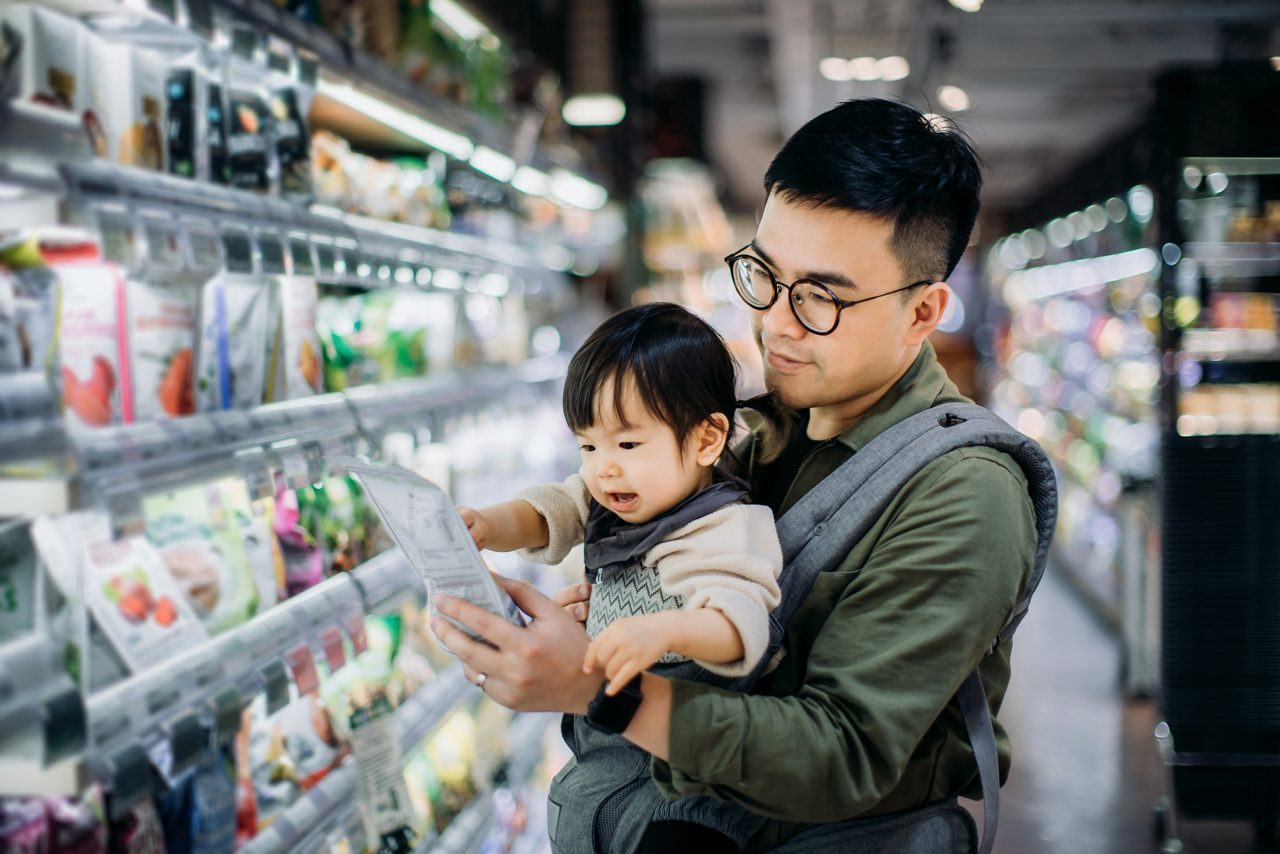 スーパーマーケットの通路で冷蔵製品を見ている男の子の赤ちゃんを持つ男性 