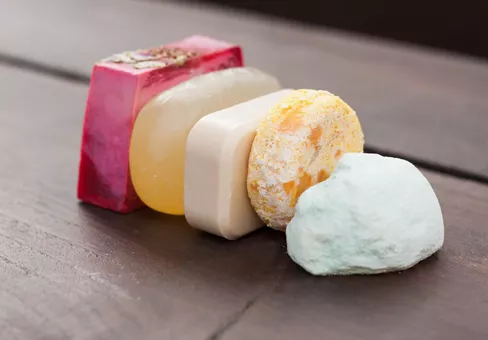 A colourful selection of stacked natural soap, shower gel, bath bomb and shampoo bars on a dark wooden table