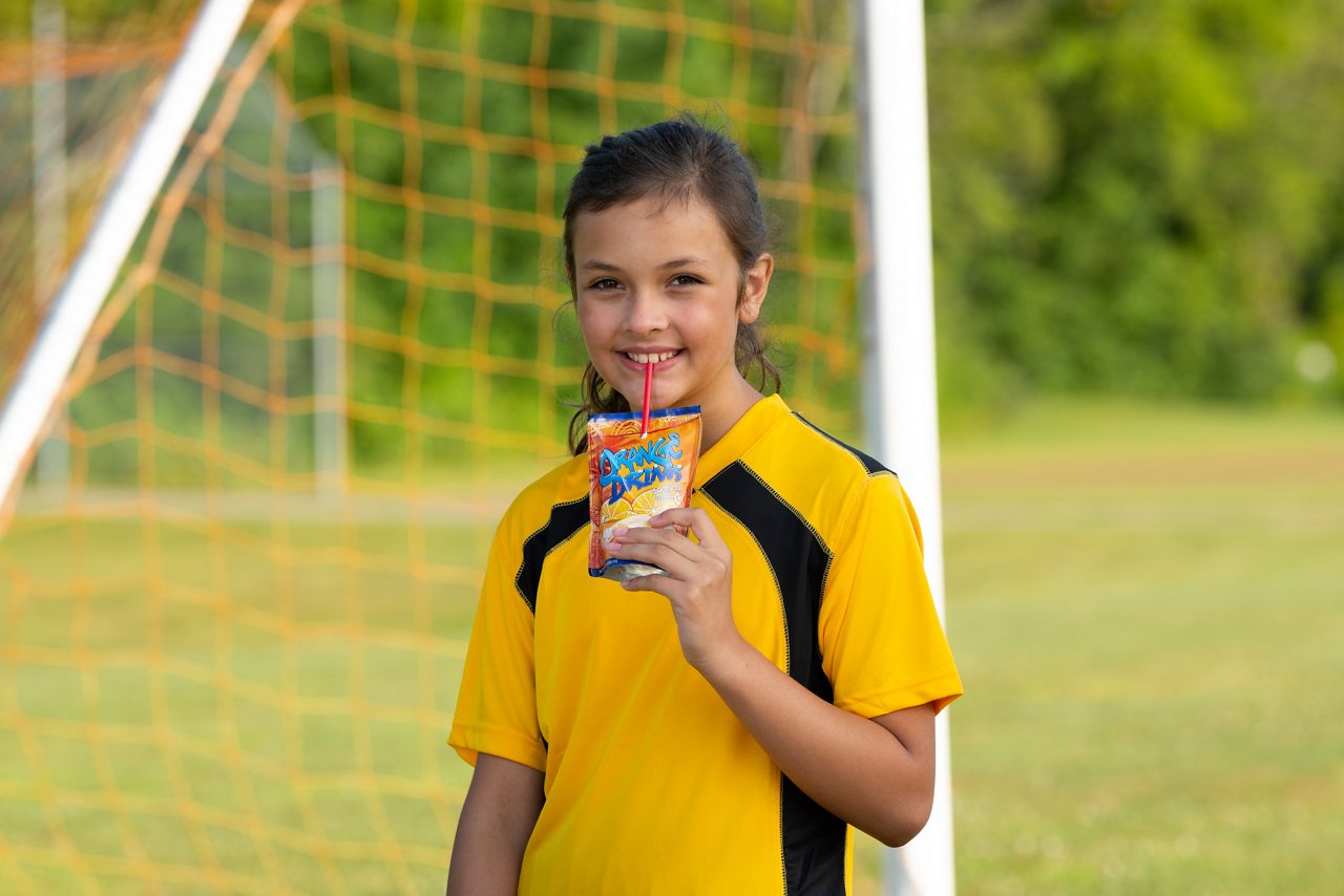 Niña de pelo oscuro con cola de caballo, con una camisa de fútbol amarilla, tomando jugo de una bolsa