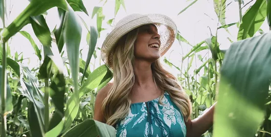 Young blonde woman wearing dress and hat walking through a corn field.
