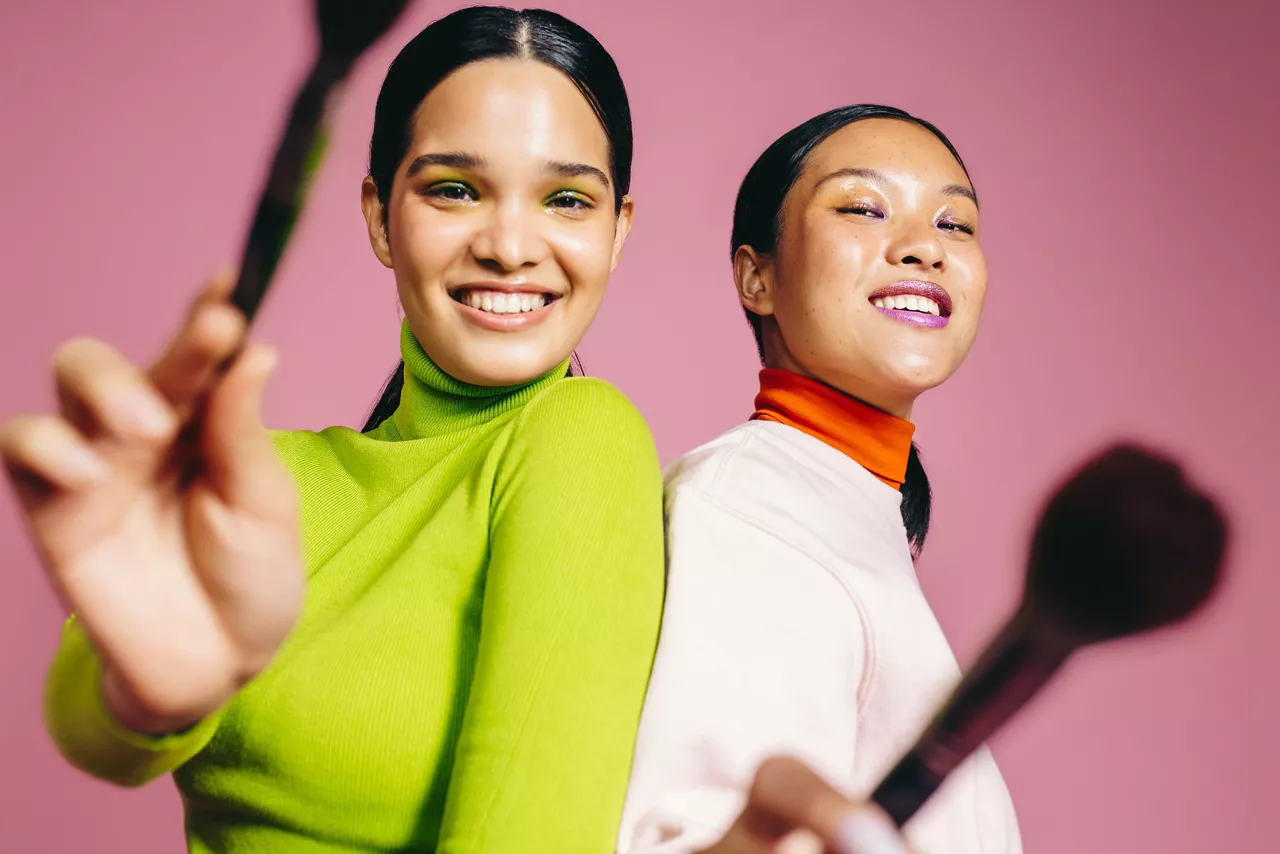 Two young women smile at the camera while holding their beauty brushes and wearing vibrant makeup. Glamorous females having fun and celebrating their love for beauty and cosmetics.