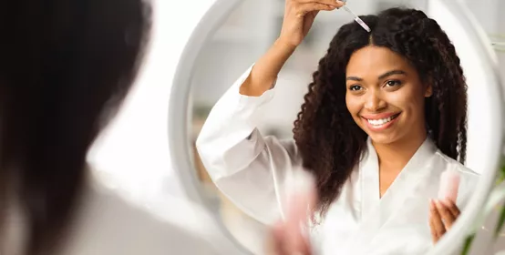 Smiling Black Woman Applying Serum For Hair Repair While Standing Near Mirror At Home, Young African American Female Making Haircare Treatment, Looking At Her Reflection, Selective Focus