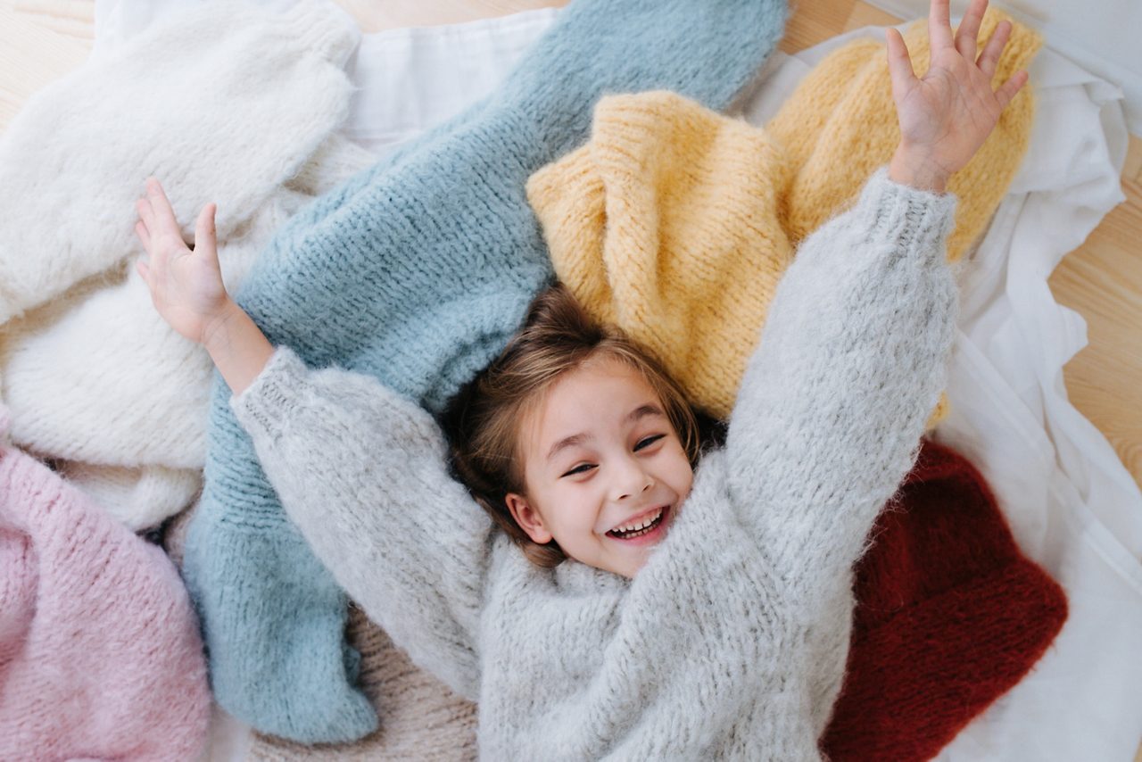 Niña estirada en el suelo, cubierta con suéteres multicolores. 