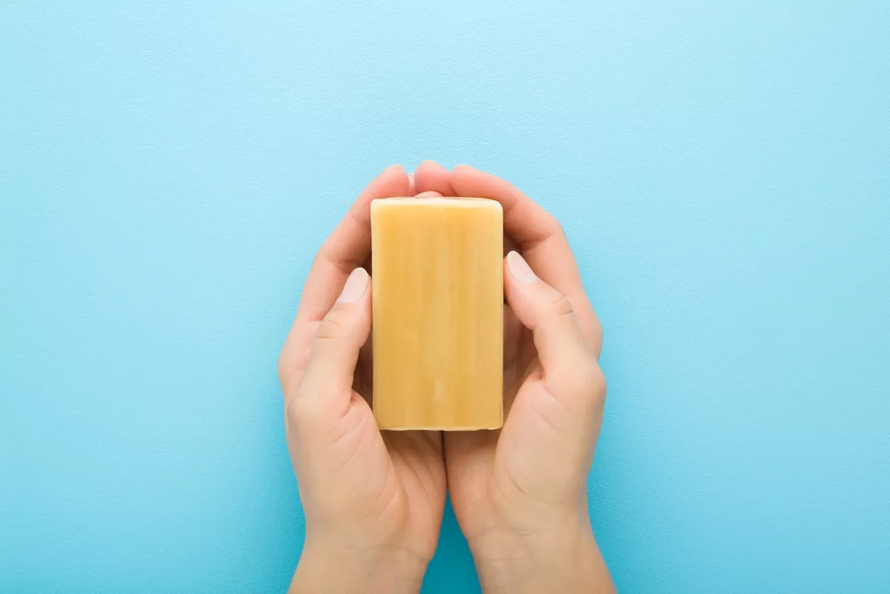 Young adult woman hands holding household soap for clothes washing on light blue table background. Pastel color. Closeup. Point of view shot. Top down view.  