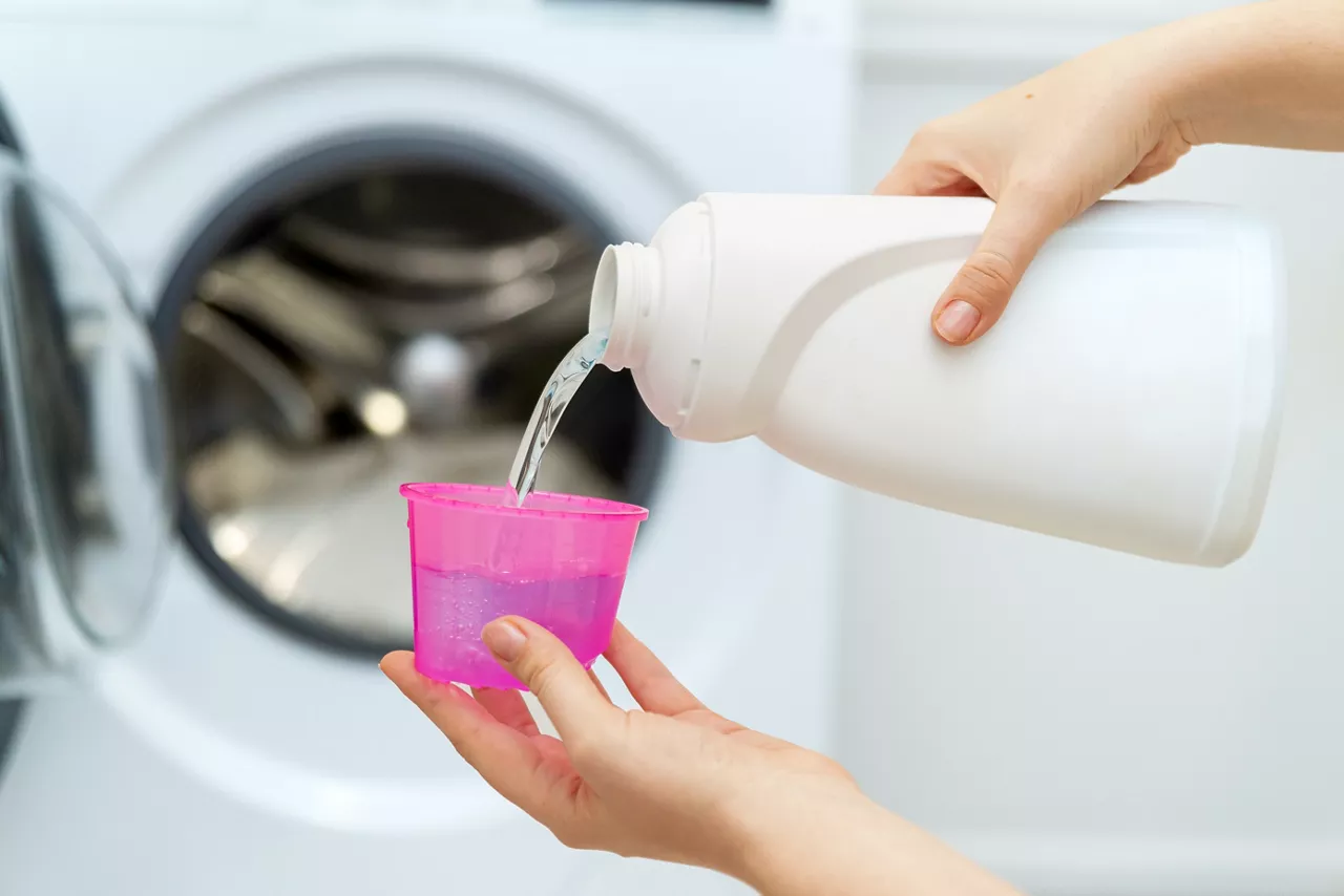 Person adding liquid laundry detergent to the washer, close up. Female hand holding laundry detergent in front of open washing machine 