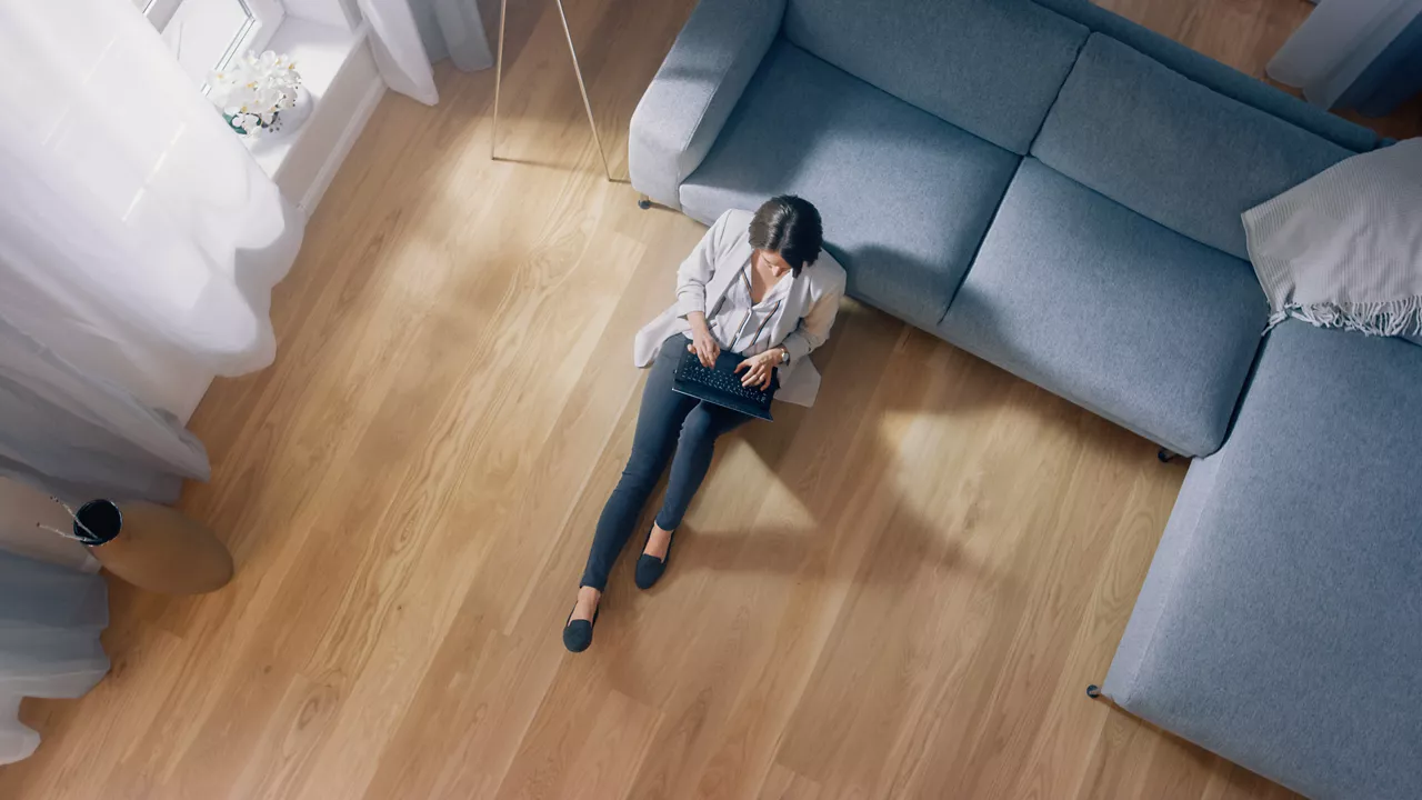Young Woman is Sitting on a Floor, Working or Studying on a Laptop. Cozy Living Room with Modern Interior, Grey Sofa and Wooden Flooring. Top View Camera Shot.
