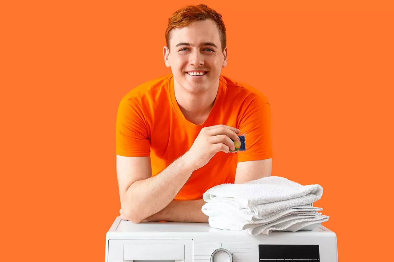 Young man with laundry capsule and towels on orange background