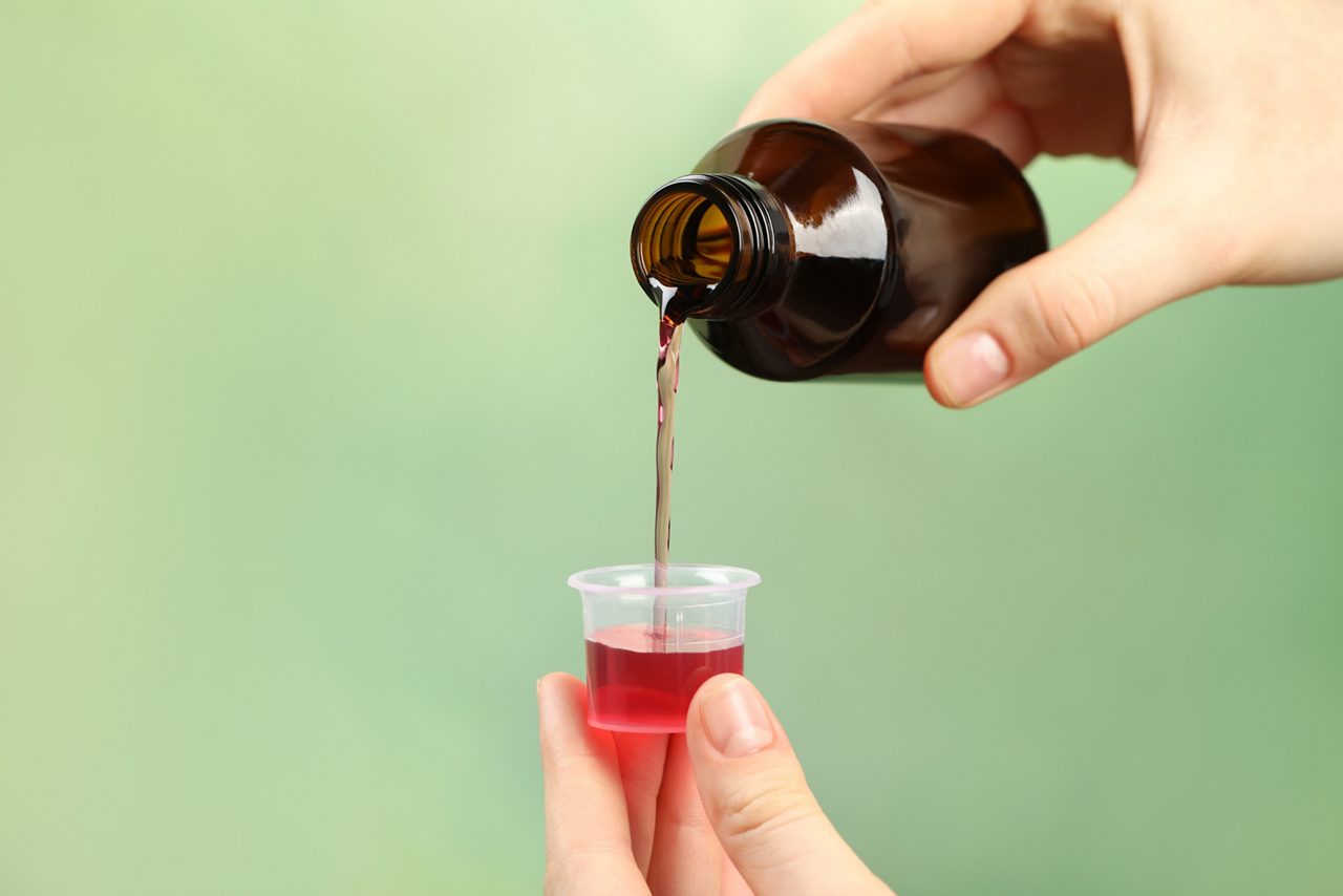 Person pouring cough syrup into measuring cup on light green background