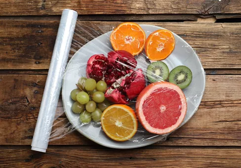 Plate of fresh fruits with plastic food wrap on wooden table, flat lay