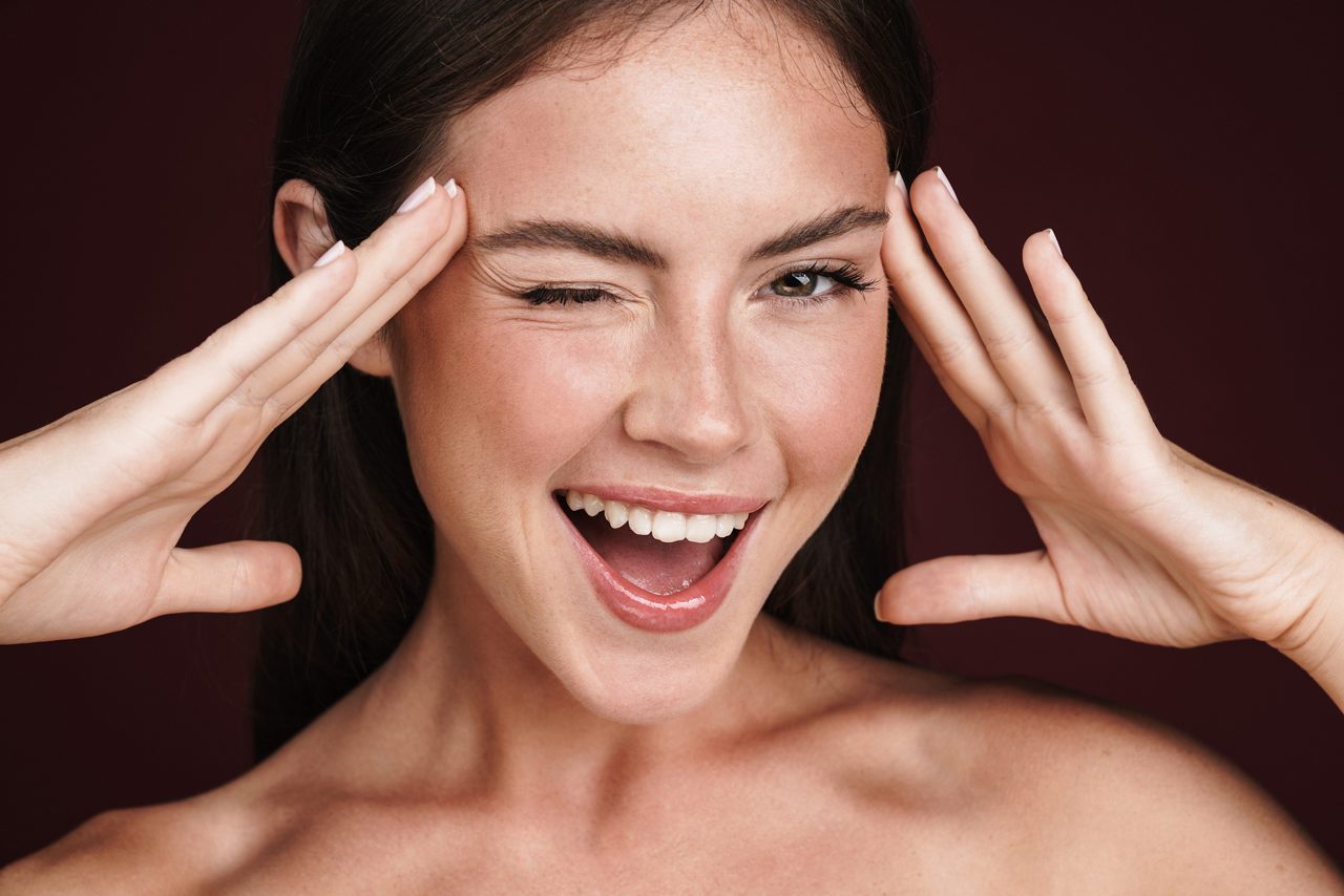 Image of smiling half-naked woman touching her temples and winking isolated over dark red background