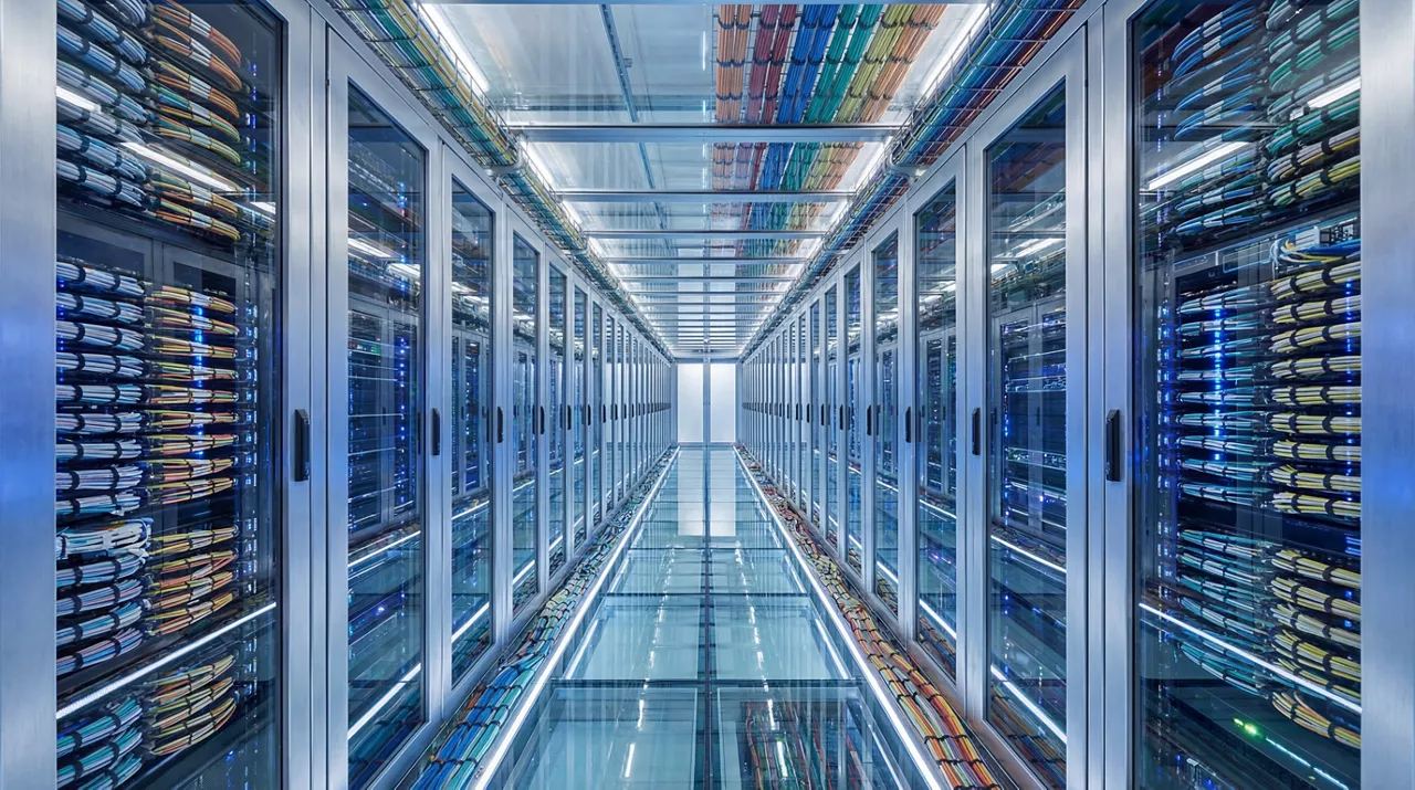 A long, brightly lit corridor in a modern data center, lined with server racks filled with equipment and cables.