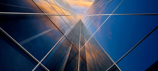 Upward perspective of a modern glass building with reflective geometric panels capturing a colorful sunset sky. The symmetrical lines converge toward the center, emphasizing height, innovation, and contemporary design.