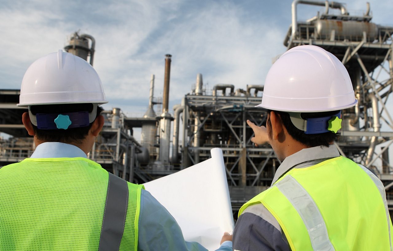Two construction employees looking at blueprints while standing in front of petrochemical machine